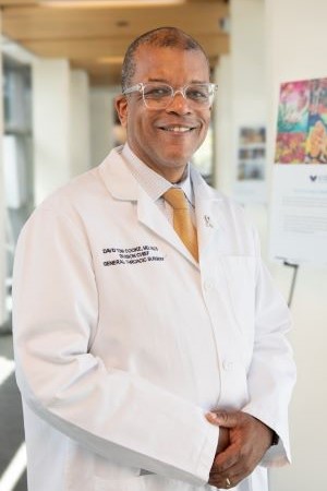 Portrait of David Cooke wearing a white lab coat and tie, standing indoors in a clinical hallway with posters and windows blurred in the background.