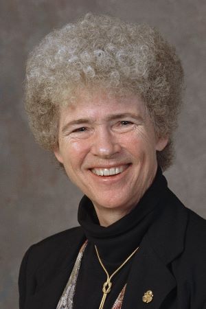 Studio portrait of Faith Fitzgerald wearing a dark jacket and necklace, photographed against a neutral textured background.