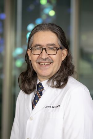 Portrait of Jorge Garcia wearing a white lab coat and tie, standing indoors with a softly blurred background.