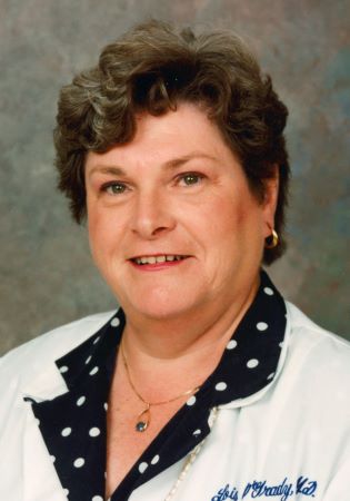 Studio portrait of Lois O’Grady wearing a white jacket over a dark polka-dot blouse, photographed against a neutral background.