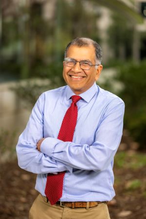 Portrait of Sharad Jain wearing a light blue button-down shirt and red tie, standing outdoors with arms crossed and campus greenery in the background.