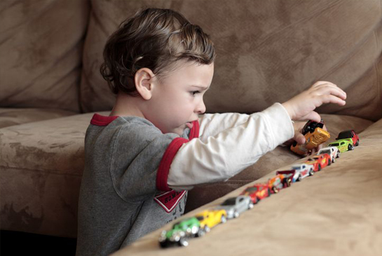 Very young male patient lines up diecast cars or hotwheels, on brown couch.