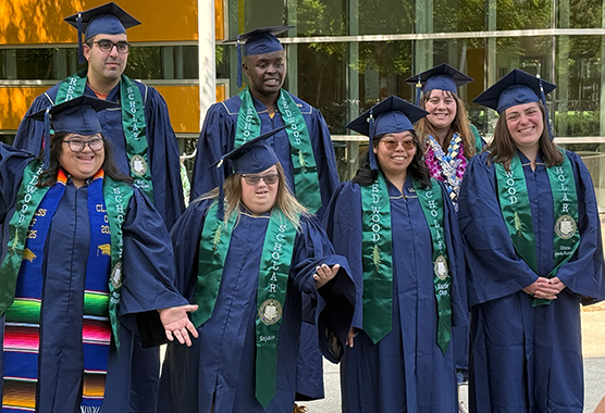  Seven Redwood SEED Scholars in navy caps and gowns pose together outside a building, wearing green stoles and colorful graduation cords