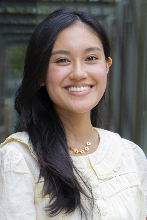 Jeramiah-Diane Bunac, smiling and standing outdoors, with long dark hair, wearing a light-colored blouse and a short necklace