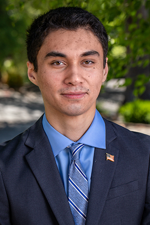 Lance Howell wearing a dark suit, blue shirt, and blue striped tie, standing outdoors with greenery in the background