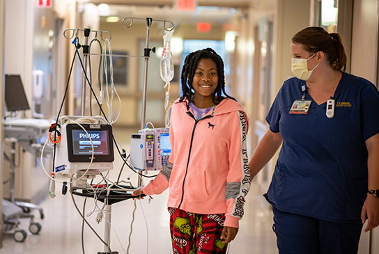 A nurse leading a girl, walking down a hallway with a computerized monitored IV cart.