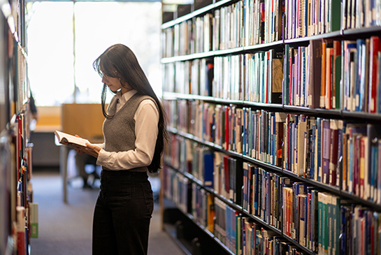 A woman standing in a row of shelves in a library looking at an open book.