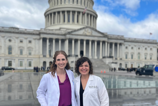 Two women smile in-front of the U.S. Capitol building wearing white medical coats.