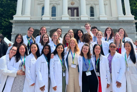 A group of residents in white coats smile and pose on steps in front of a building with large columns above a stone wall.