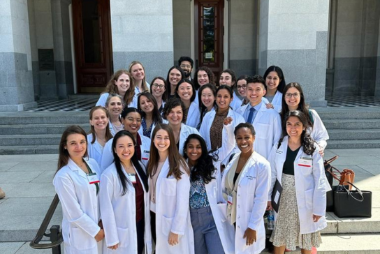 A group of residents in white coats smile and pose on steps in front of a building with large columns in-front of the door.