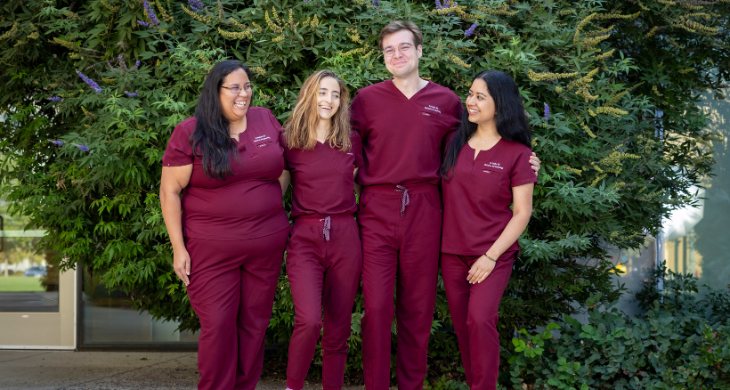 2027 class of OBGYN residents smiling near trees and bushes.