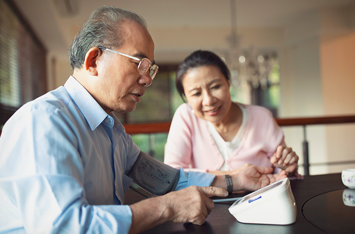 Elderly man sitting at kitchen table checking blood pressure, wife looking on