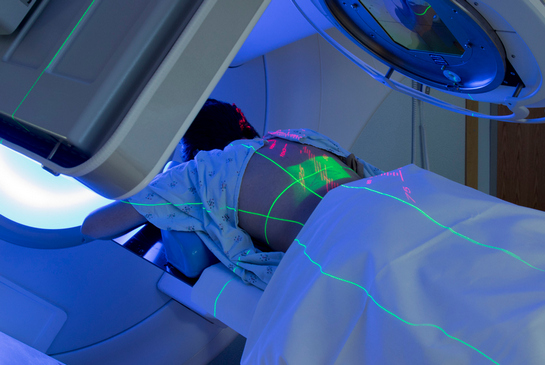 A patient positioned face down on a treatment table under a medical device, with glowing green and red guide lines projected across patient’s back.
