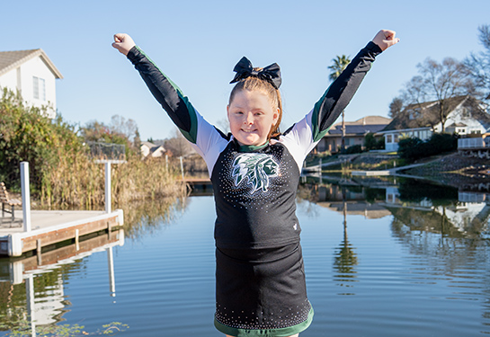 A young girl stands on a deck with water behind her. She wears a cheerleader uniform and stands with arms in a “Y” shape.