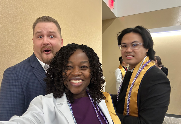 Three graduates smile for a celebratory selfie at a commencement event, wearing white coats, honor cords and graduation regalia.