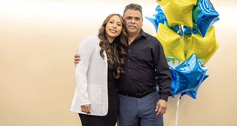 Two people embrace and smile for a photo during a white coat or recognition ceremony, standing beside blue and gold star-shaped balloons.