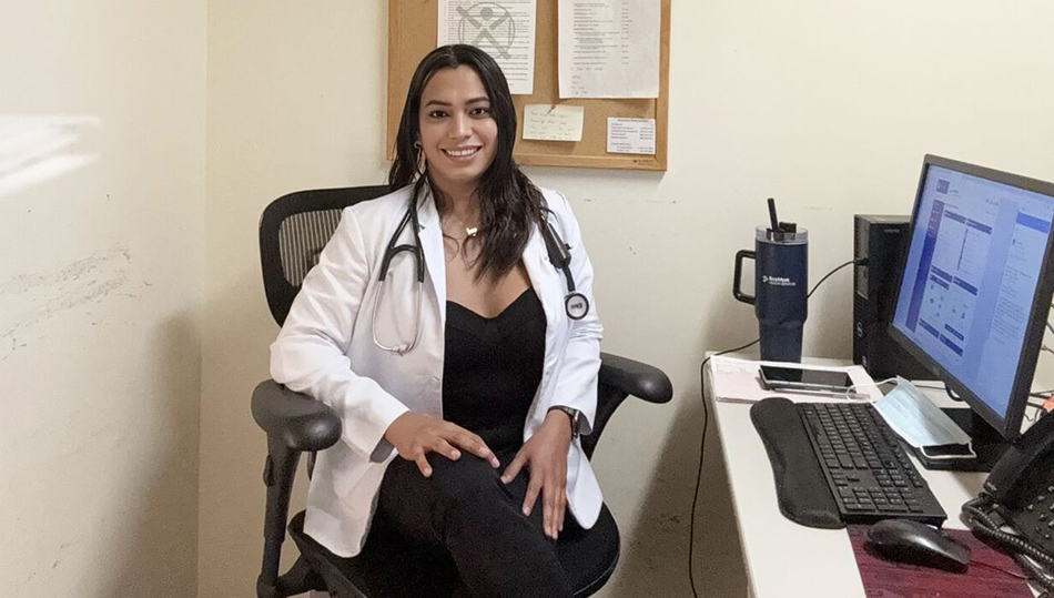 Nurse wearing a white coat and stethoscope sits at a desk in a clinical office, smiling toward the camera with a computer workstation nearby.