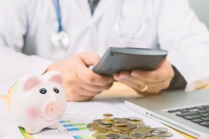 person in lab coat sitting at laptop, using calculator with piggy bank and money on table