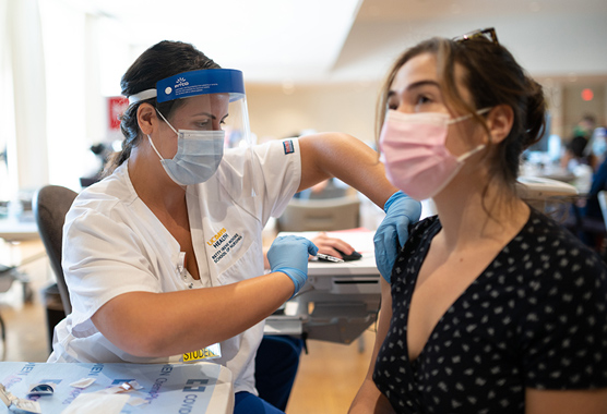 Nurse wearing a mask and face shield administering a vaccine to a female patient.